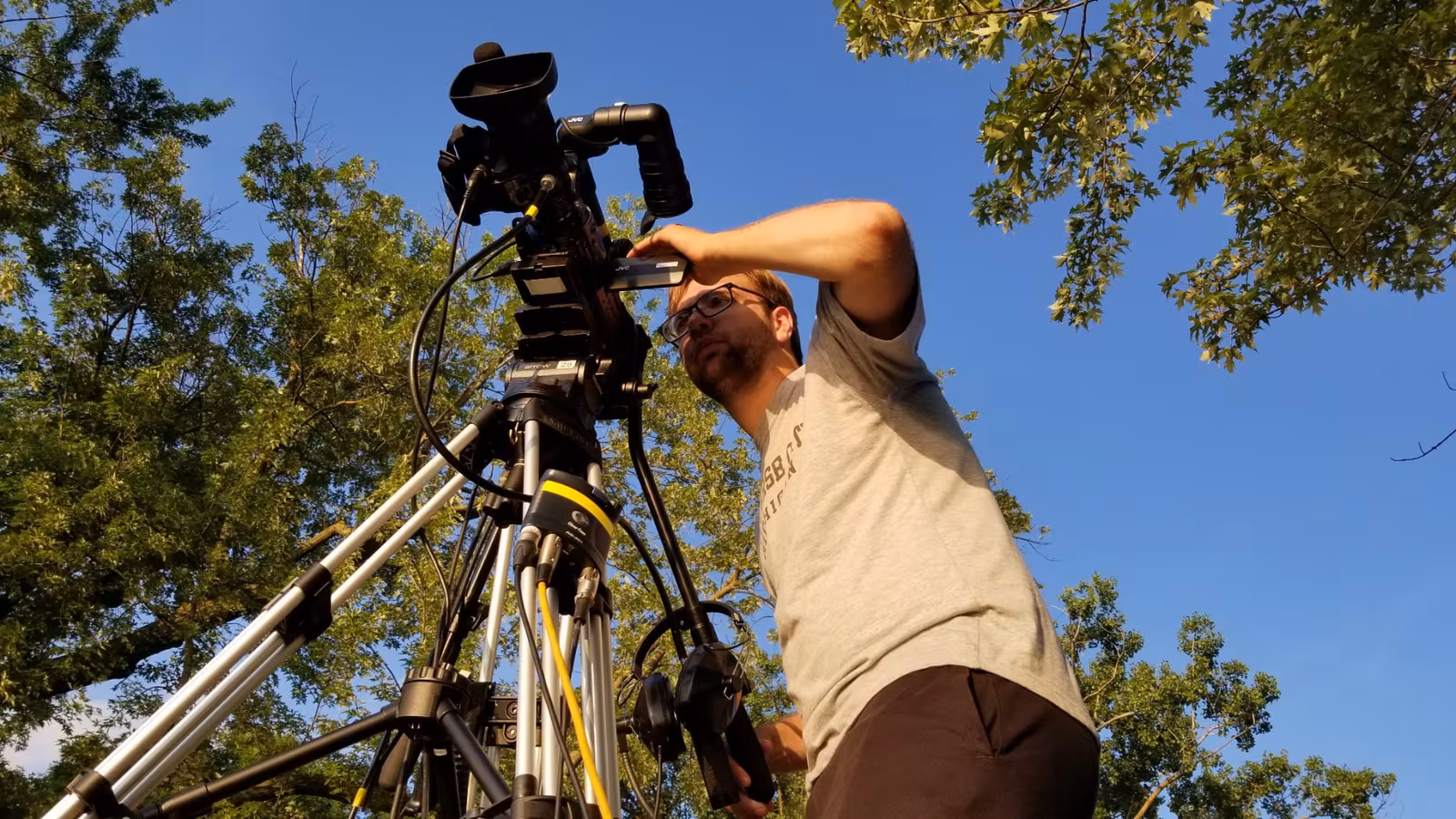 Videographer operating a broadcast camera on a tripod outdoors beneath a canopy of trees