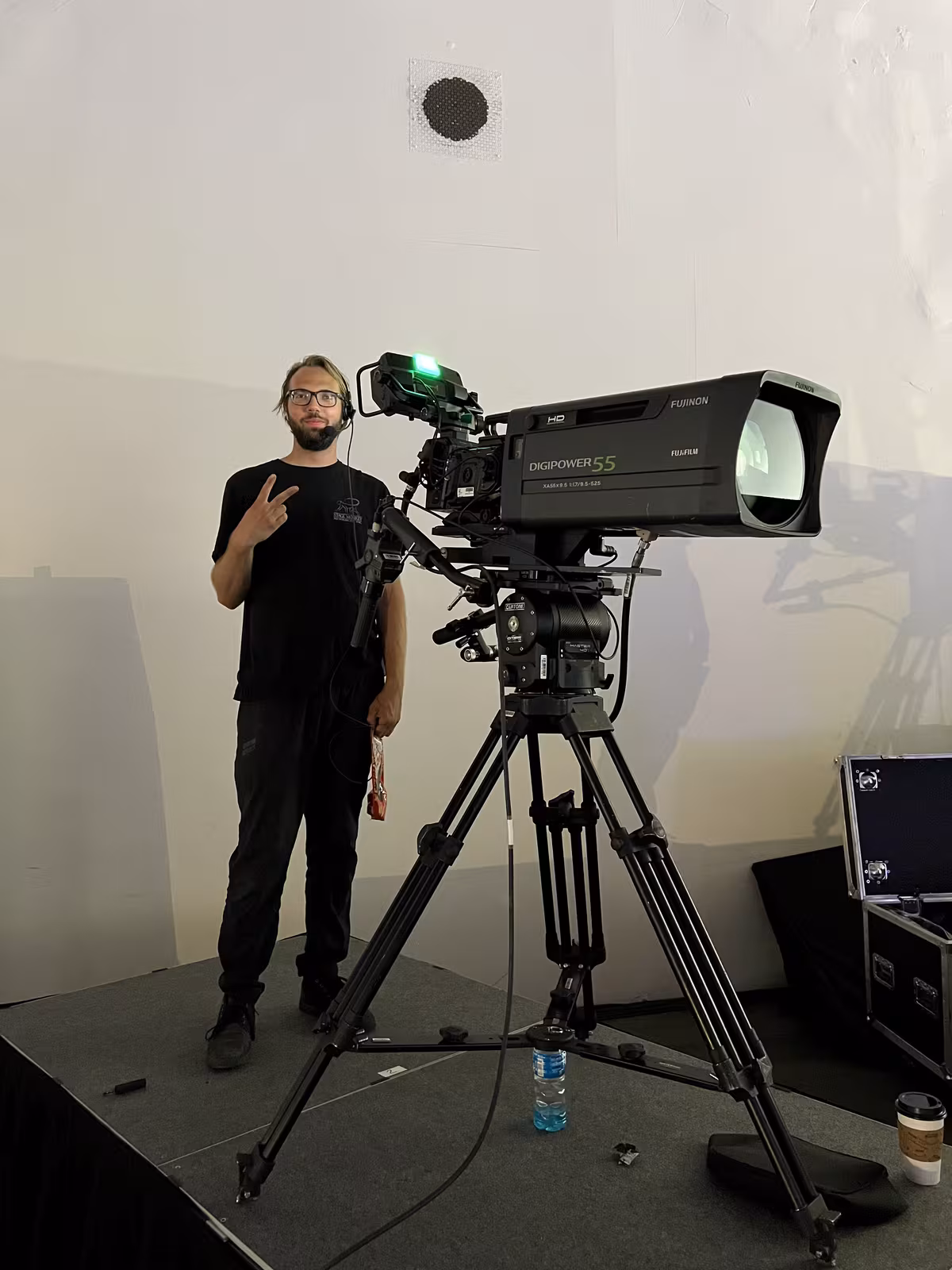 Camera operator standing beside a broadcast camera with a long telephoto lens on a stage