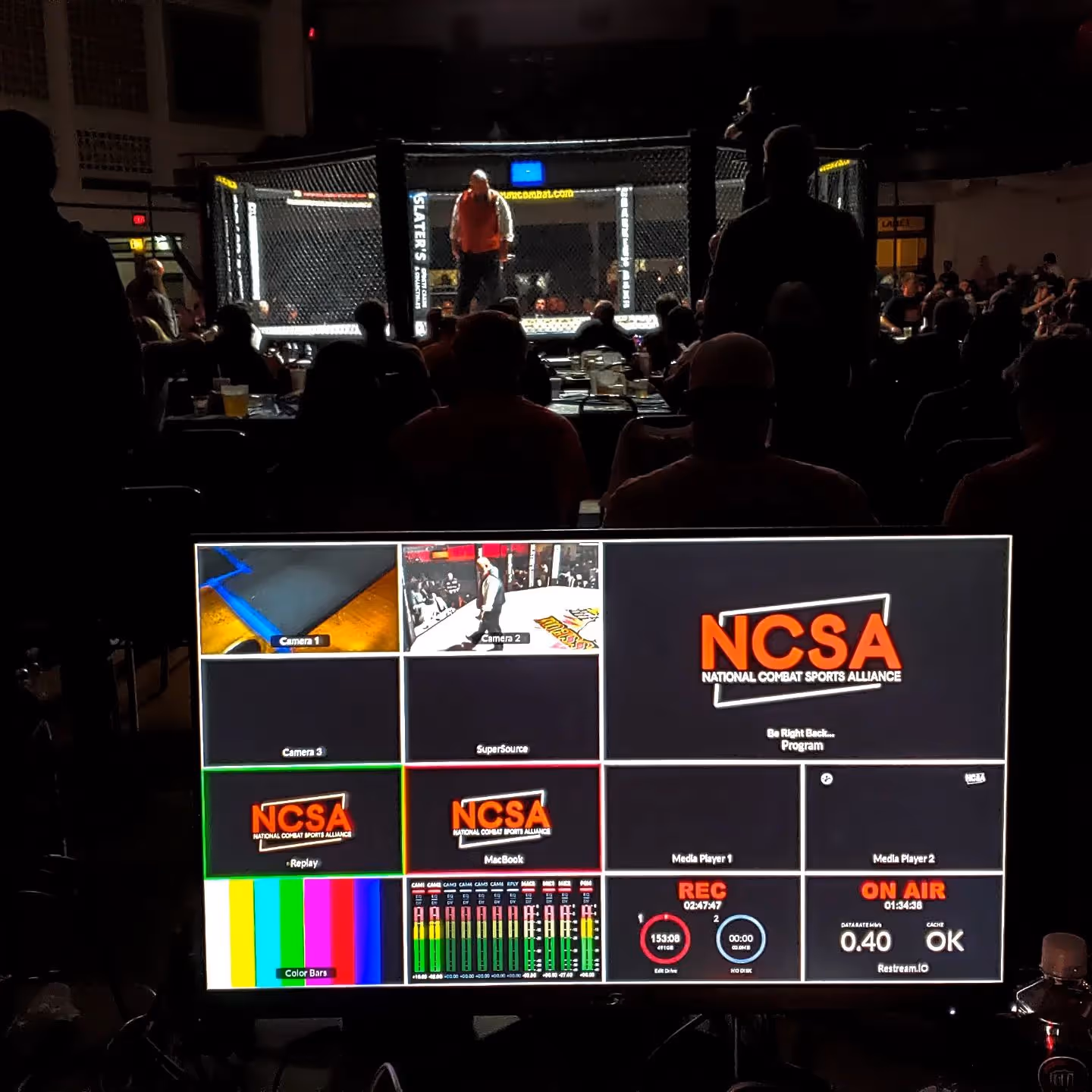View of a mixed martial arts cage match with silhouettes of audience members and a control monitor