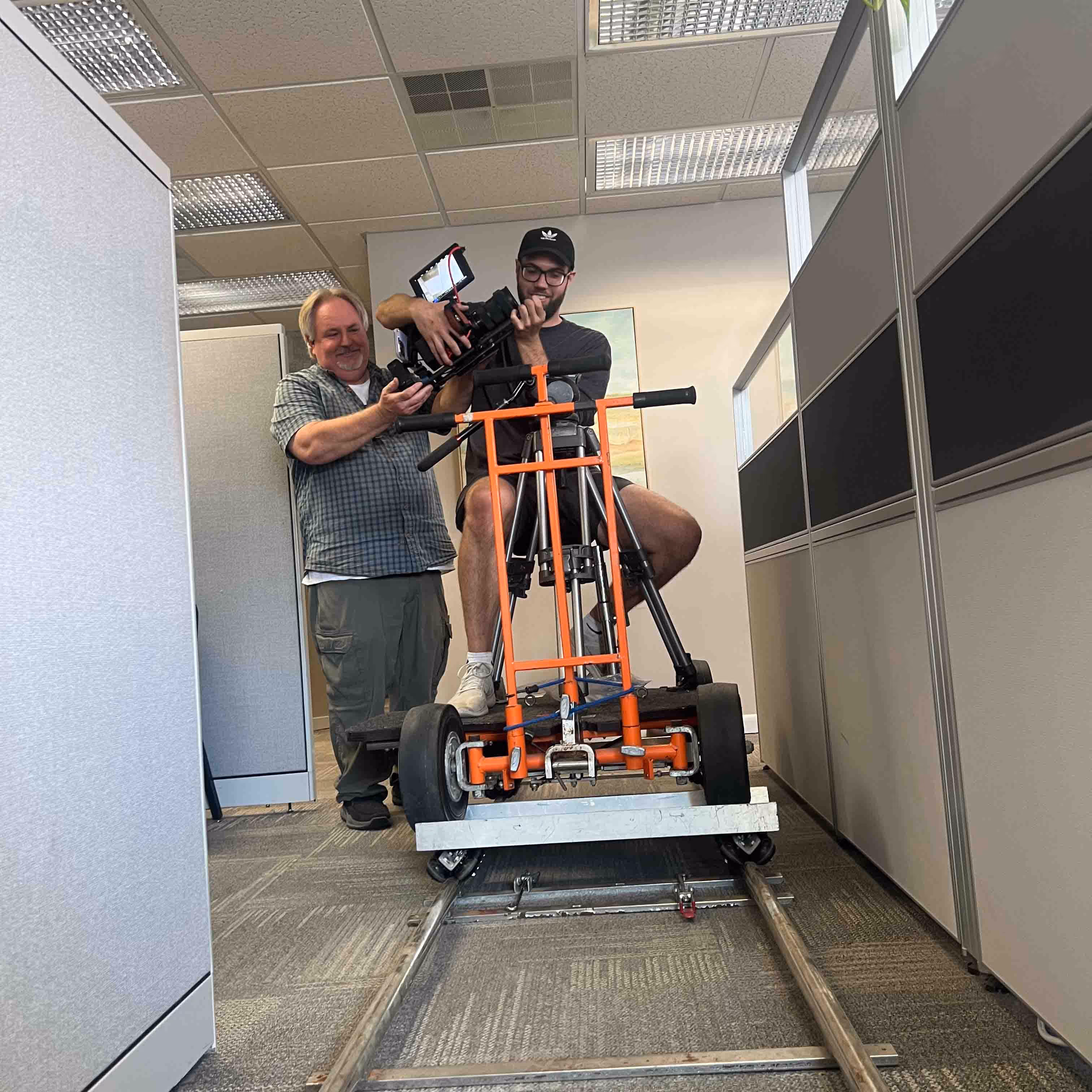 A man operating a camera rig mounted on a dolly on track in an office hallway