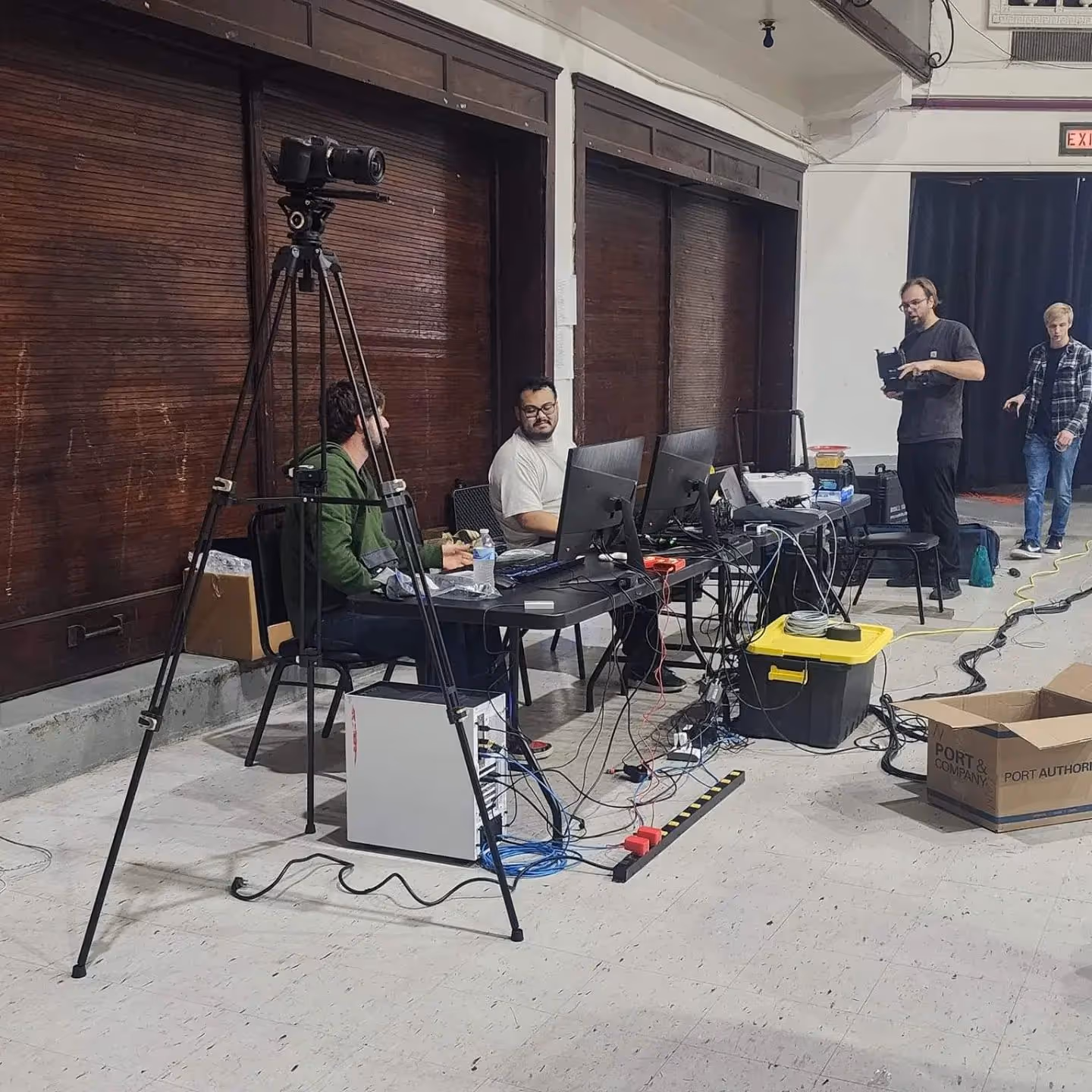 Two men working at computers with cables and equipment, camera on tripod in foreground