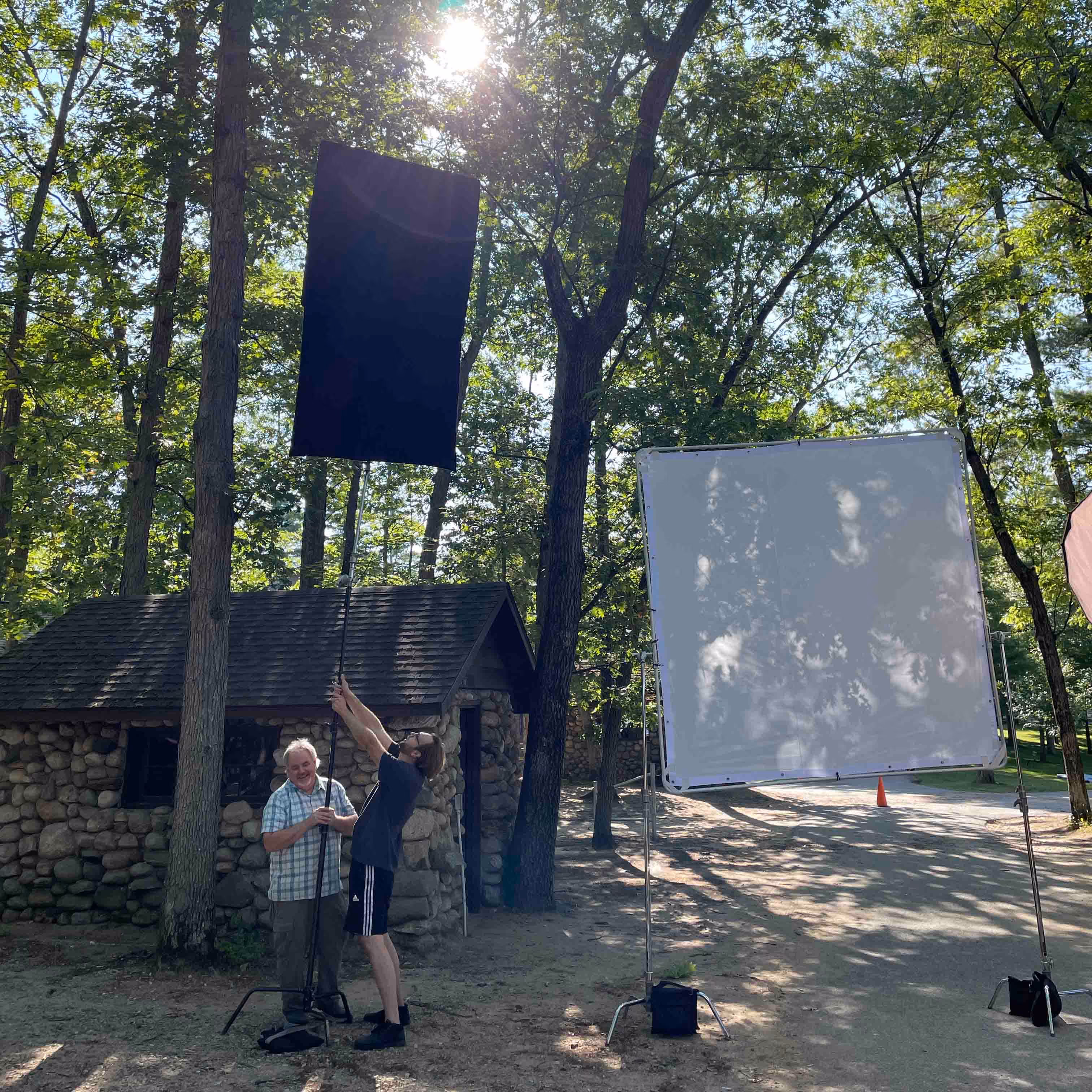 Two men adjusting fabric on a pole outdoors near a stone cabin surrounded by trees